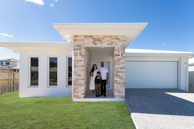 Family of three standing in front of a modern white house with stone accents, blue sky.