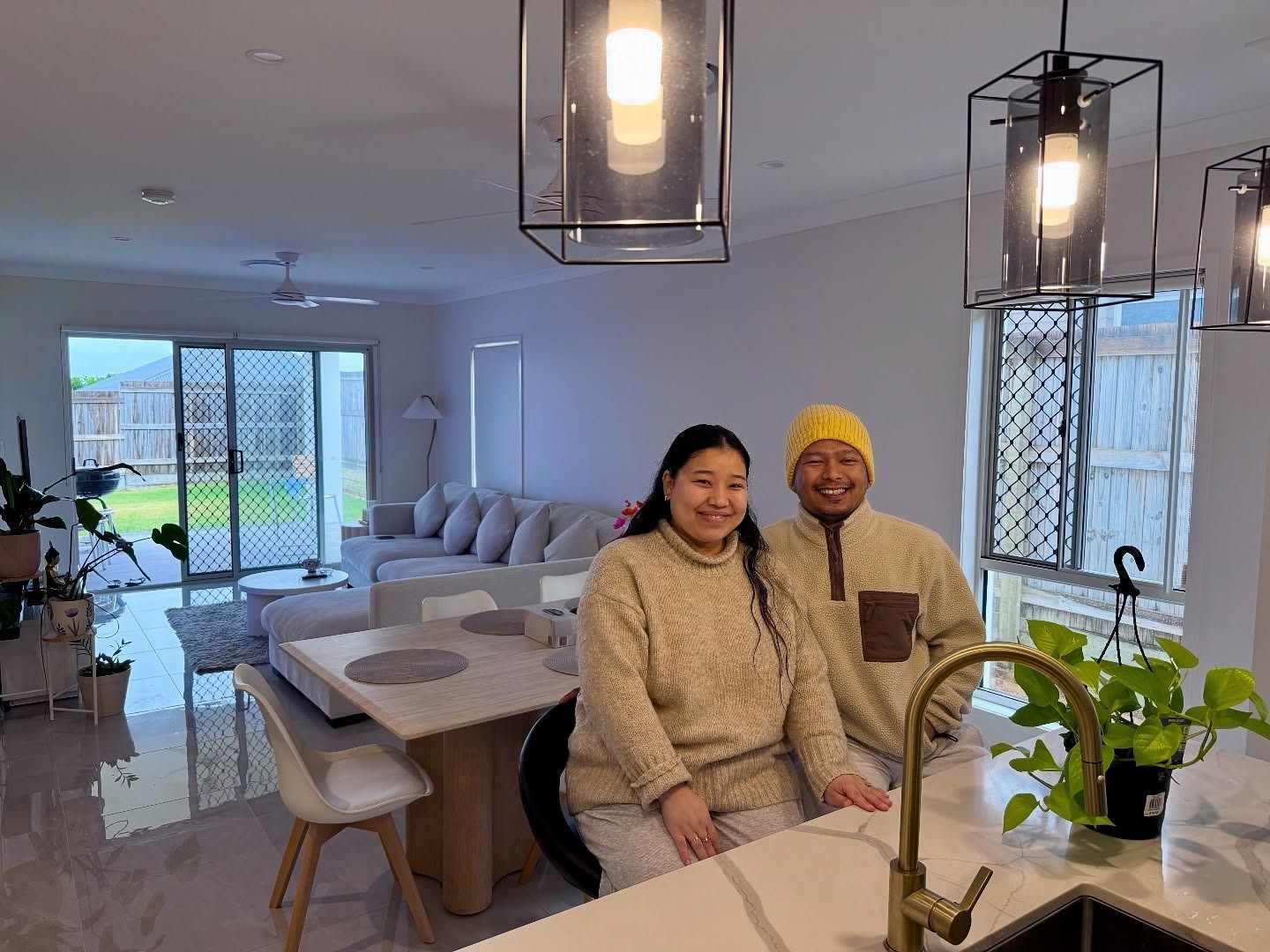 A couple smiles in their new kitchen, wearing matching beige sweaters. Modern home interior, open plan.