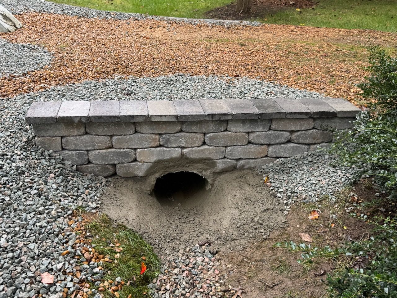 Stone-covered culvert bridge with an opening for water flow, surrounded by gravel and foliage.