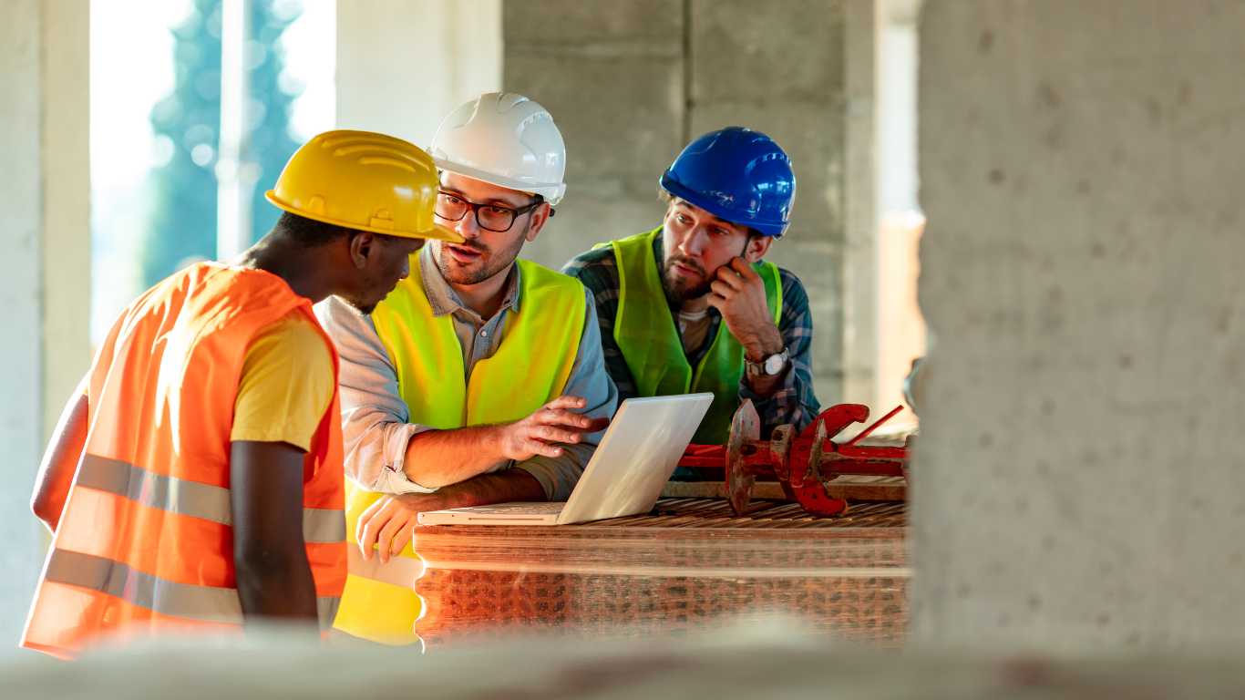 A group of construction workers are looking at a laptop on a construction site.