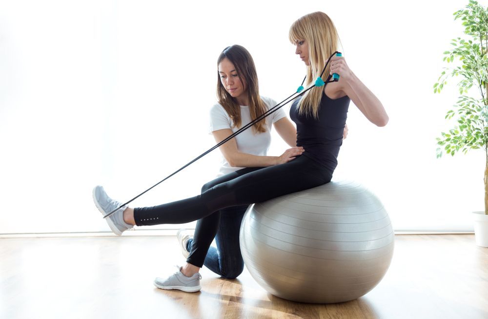 Woman on exercise ball uses resistance band, guided by a trainer. Indoors, natural light. — Bourbong Street Physiotherapy Centre in Bundaberg West, QLD