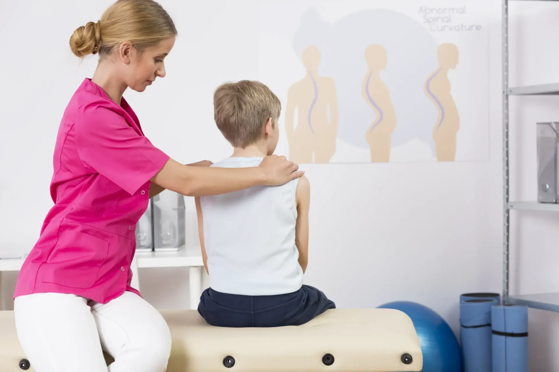 Doctor examines a child's back for scoliosis, seated on an examination table. A poster with spine curves is visible. — Bourbong Street Physiotherapy Centre in Bundaberg West, QLD