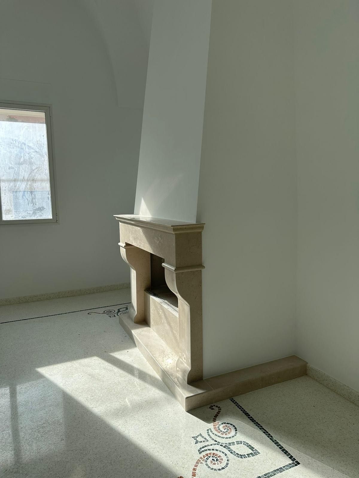 In una stanza con pavimento in piastrelle chiare screziate e decorate con motivi a mosaico, un camino in pietra è addossato a una parete bianca.