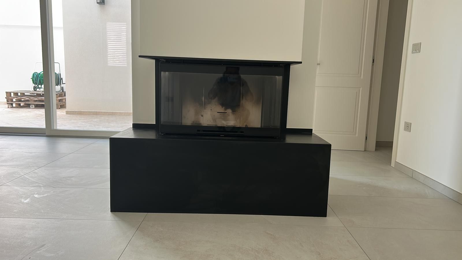 In una stanza con pavimento in piastrelle chiare, un moderno inserto per camino nero è posizionato su una base rettangolare nera, addossato a una parete bianca.