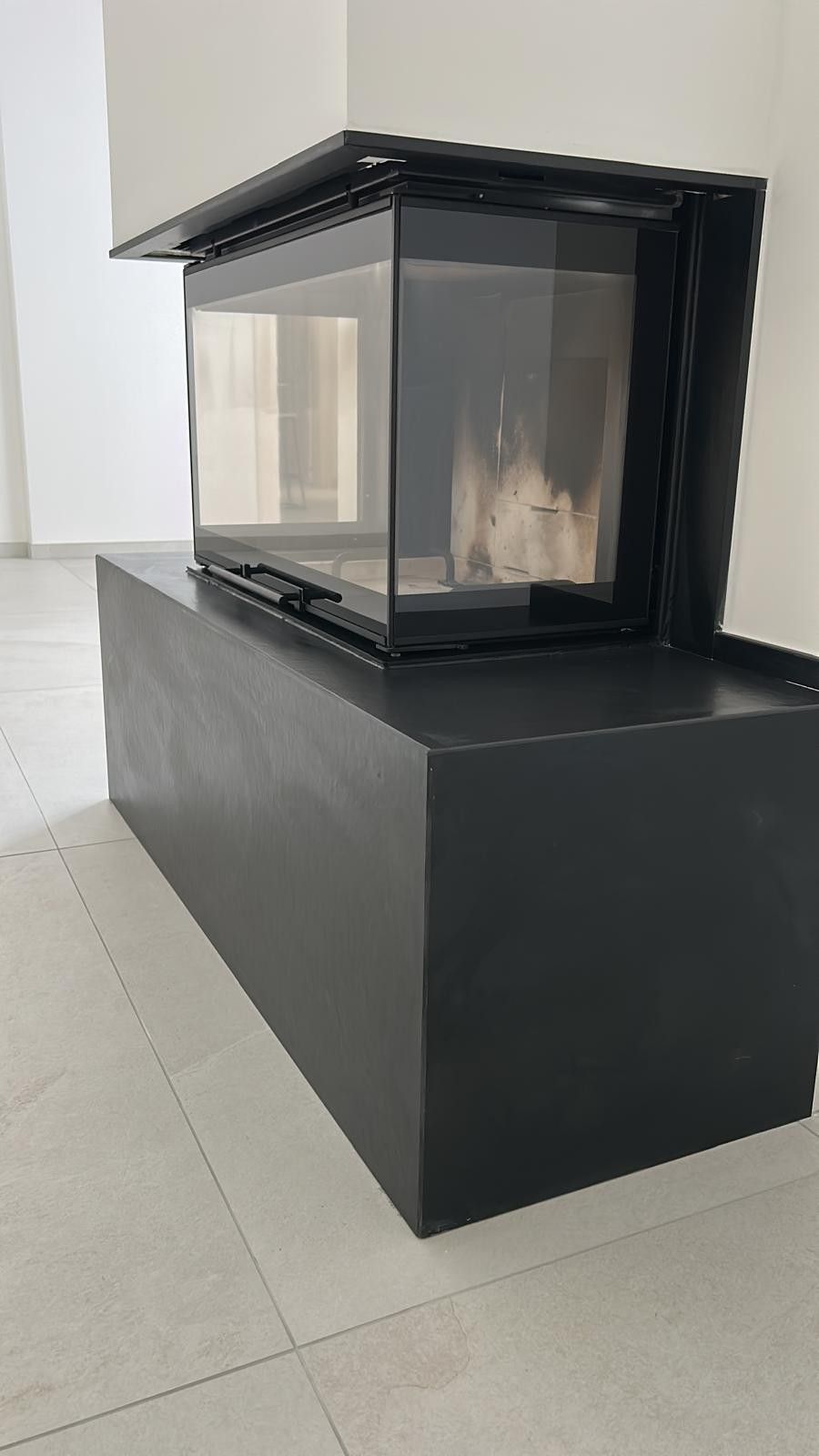 Un moderno inserto per camino nero è posizionato su una base rettangolare nera, addossato a una parete bianca su un pavimento piastrellato.