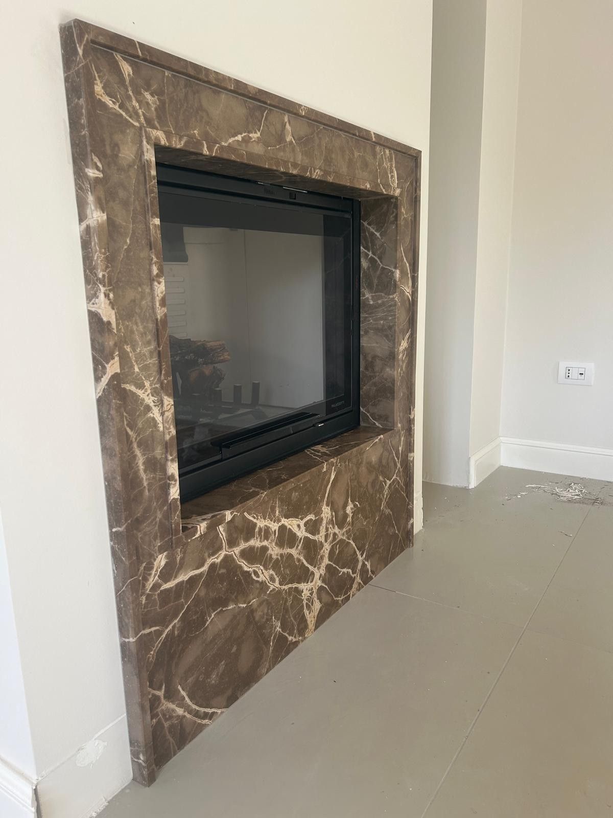 Una moderna cornice per camino in marmo marrone scuro con venature bianche, installata su una parete bianca.