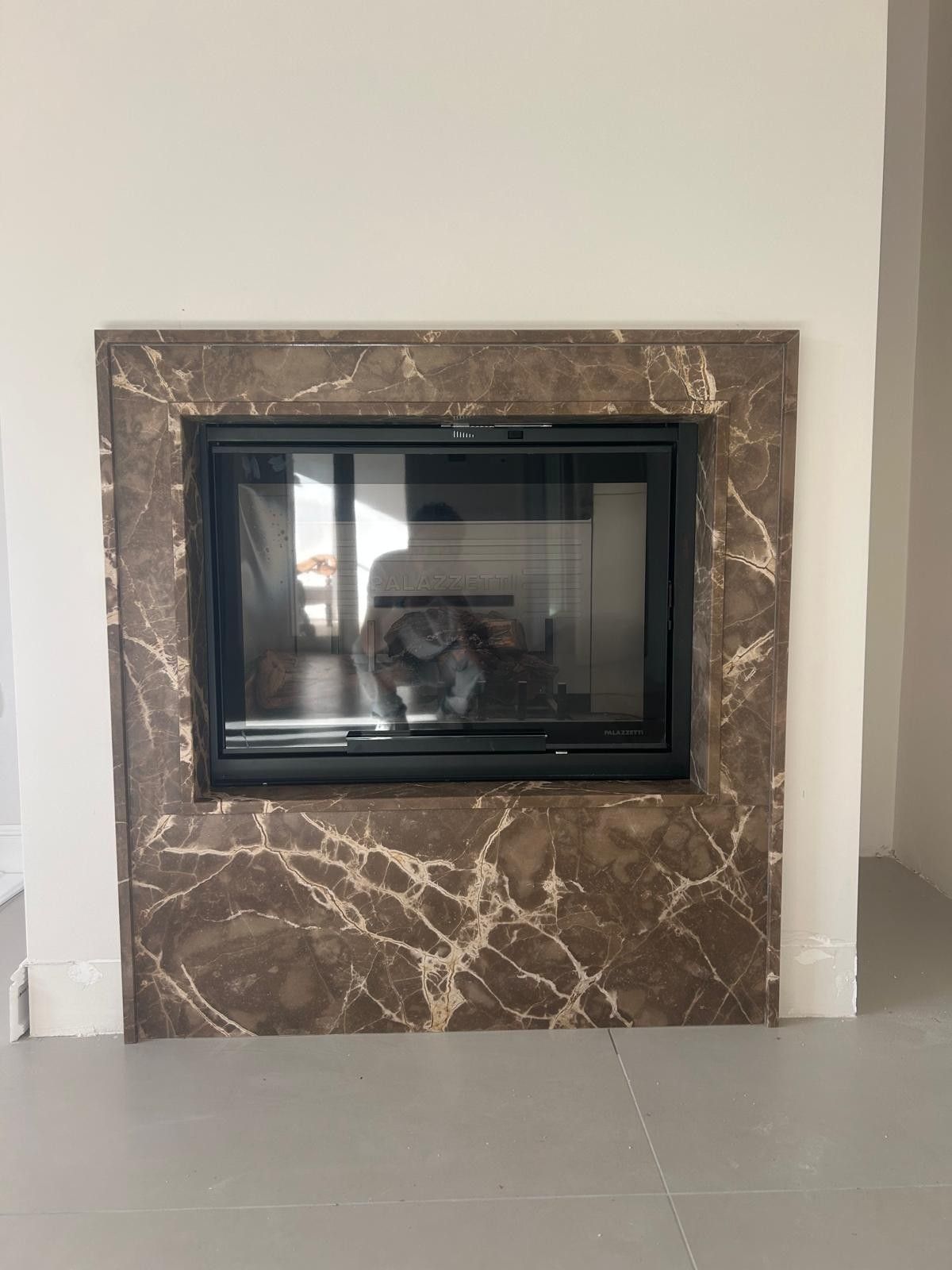Un camino con cornice in marmo marrone venato di bianco, incastonato in una parete bianca sopra un pavimento grigio.