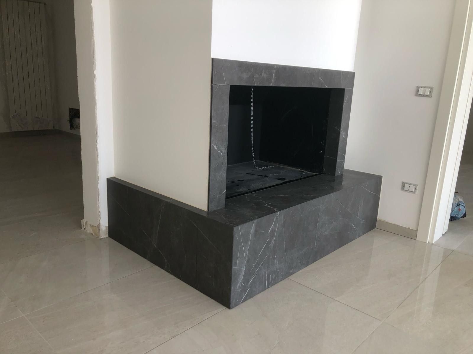 Camino moderno con cornice e focolare in marmo grigio scuro, inserito in una stanza con pavimento in piastrelle di colore chiaro.