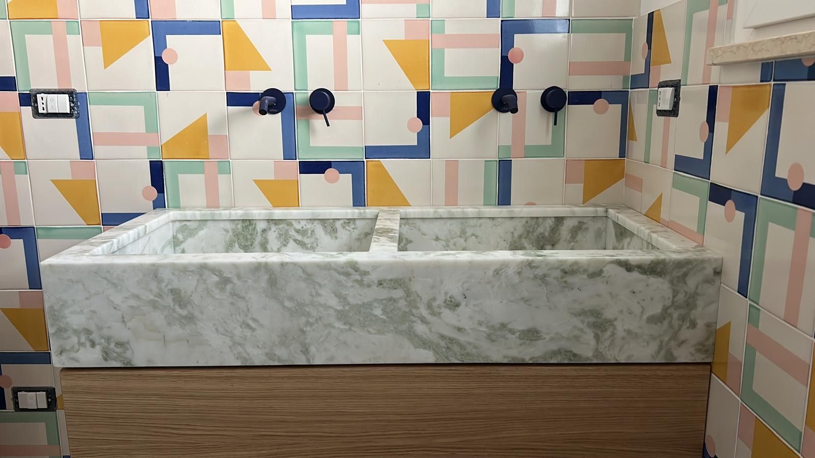 Un doppio lavabo in marmo incastonato in una parete rivestita di piastrelle con motivi geometrici multicolori.