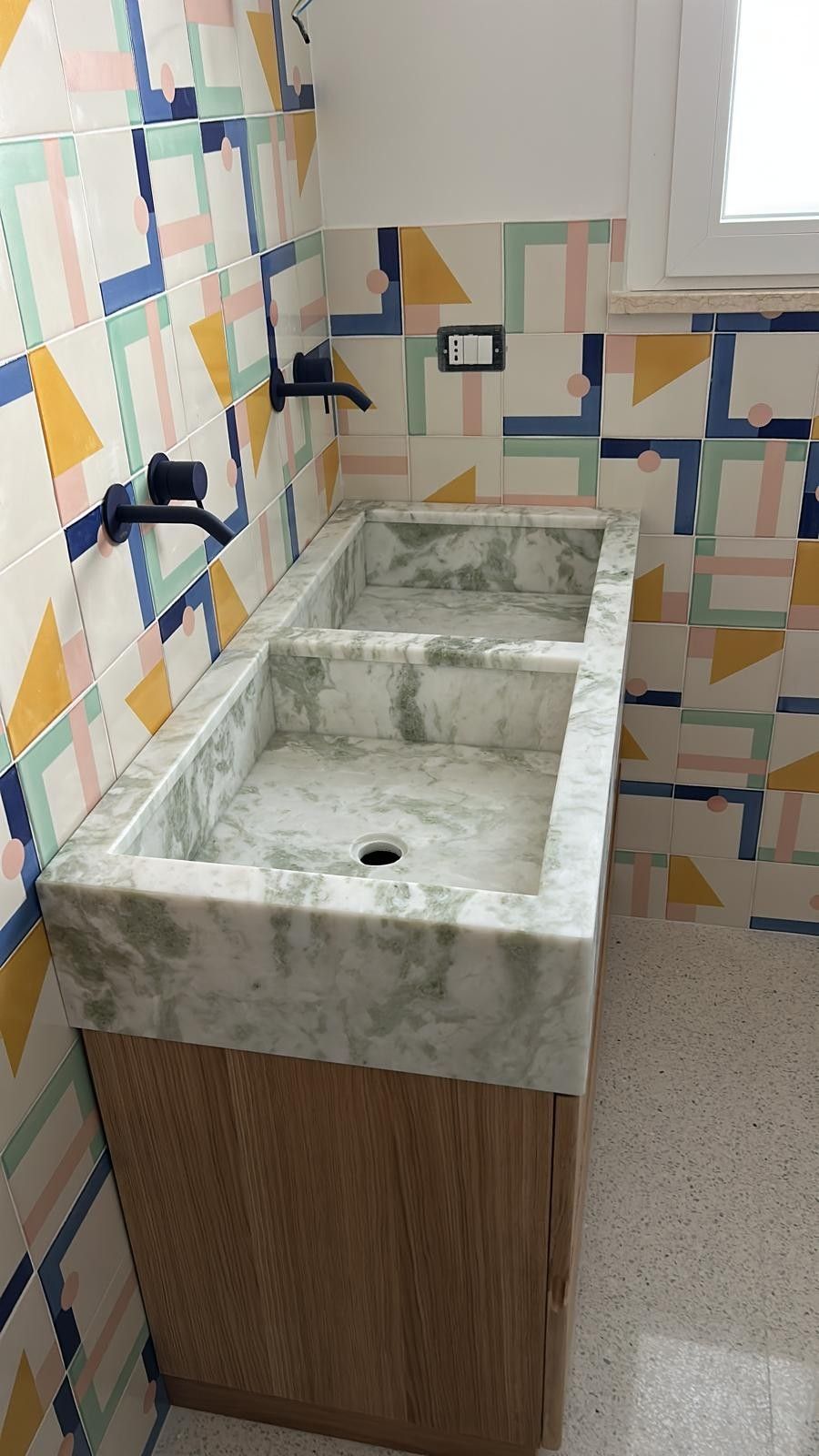 Un doppio lavabo in marmo con rubinetteria nera opaca, montato su una parete piastrellata con motivi decorativi, sopra un mobiletto in legno chiaro.