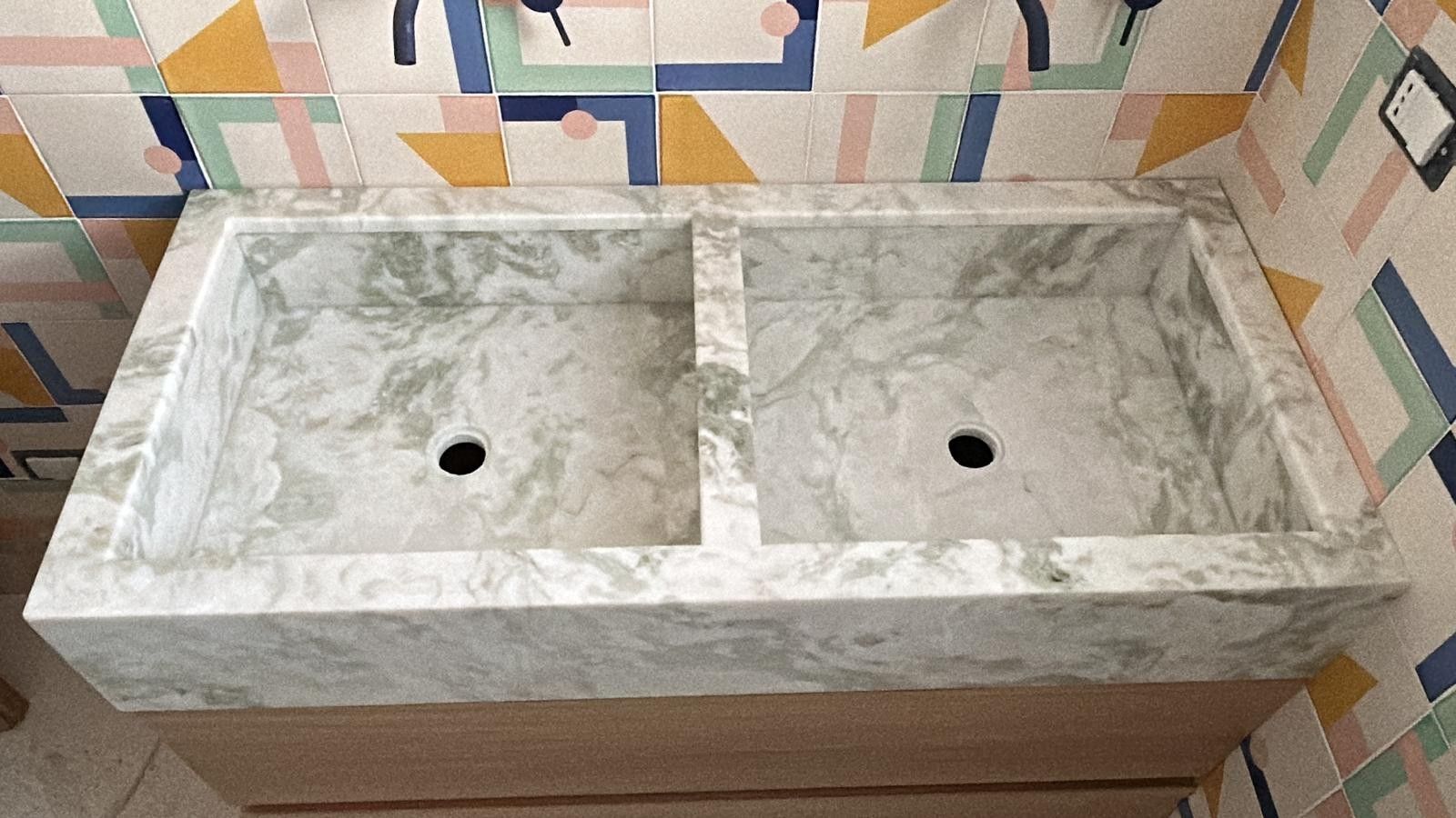 Un moderno lavabo a doppia vasca in pietra con venature di marmo grigio chiaro, montato su un mobiletto in legno e rivestito con piastrelle geometriche.