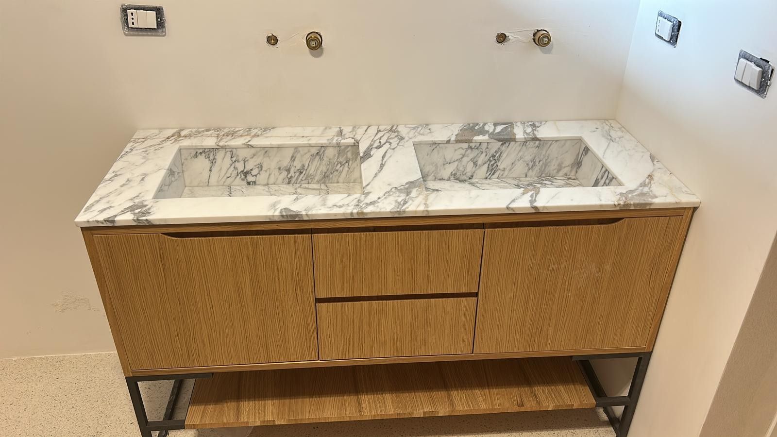 Doppio lavabo con piano in marmo e mobili in legno chiaro, incastonato in una parete bianca con rubinetteria a parete.