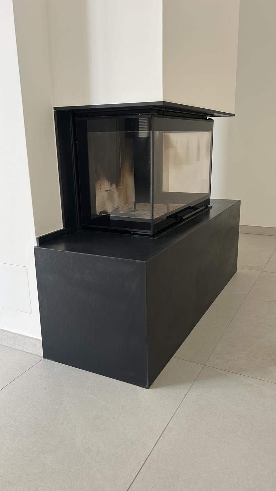 In una stanza con pareti bianche e pavimento piastrellato chiaro, un inserto per camino rettangolare di colore nero poggia su un piedistallo nero abbinato.