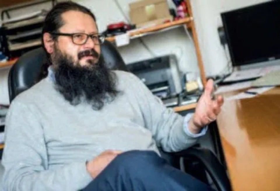 Uomo con occhiali e barba, che gesticola mentre è seduto su una sedia da ufficio, vicino a una scrivania.