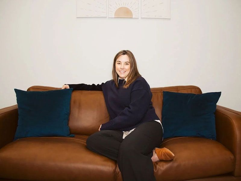 Person sitting on a brown leather couch between two dark blue pillows, smiling in a bright room.