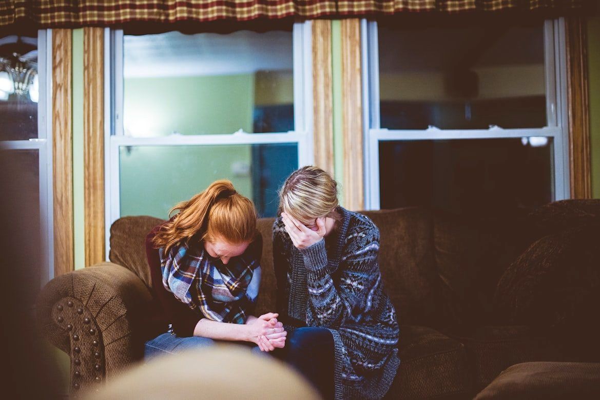Two people sit on a couch, leaning over a small object in their hands by a window at night.