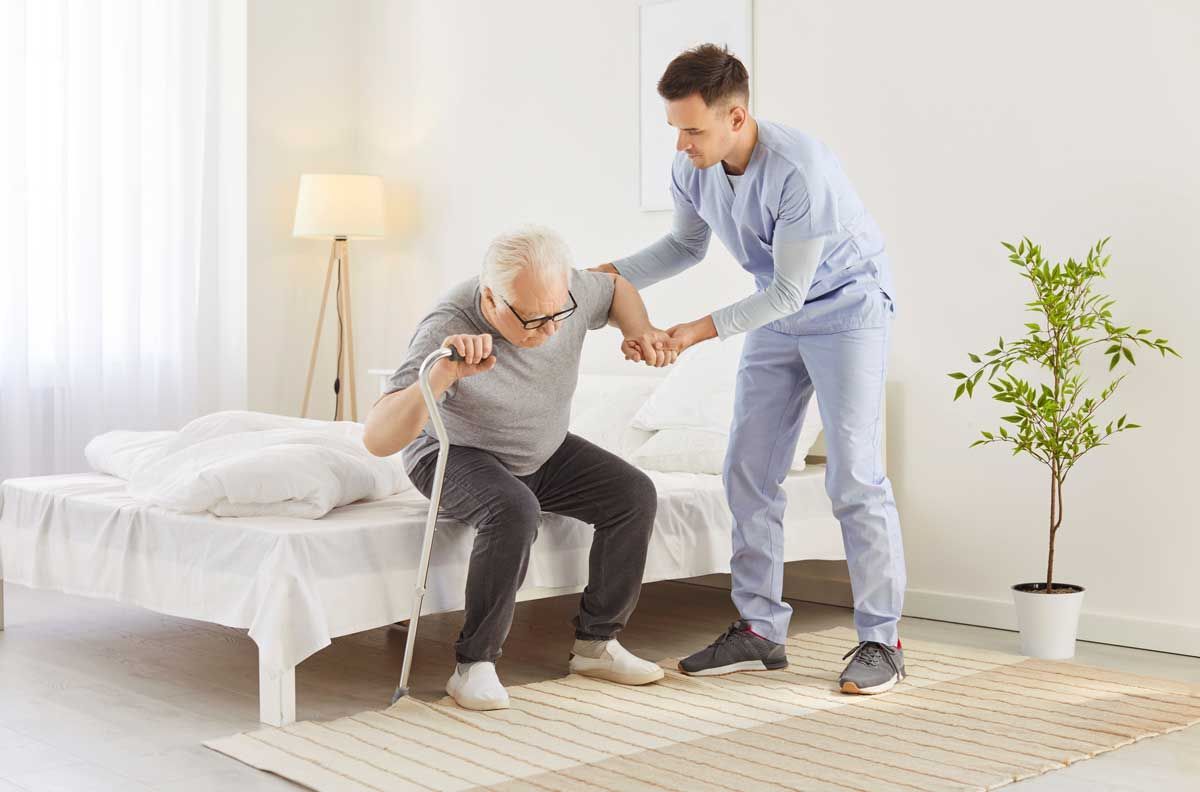 A healthcare worker assists an elderly man with a cane getting out of bed.