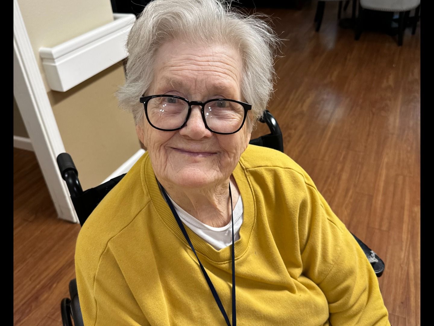 Elderly woman in a wheelchair, smiling, wearing glasses and a yellow sweater. Indoors.