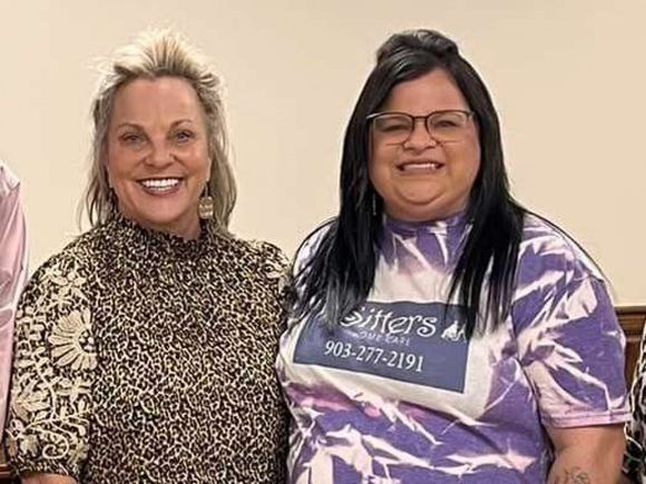 Two women smiling, indoors; one in animal print, the other in a tie-dye shirt with business logo.