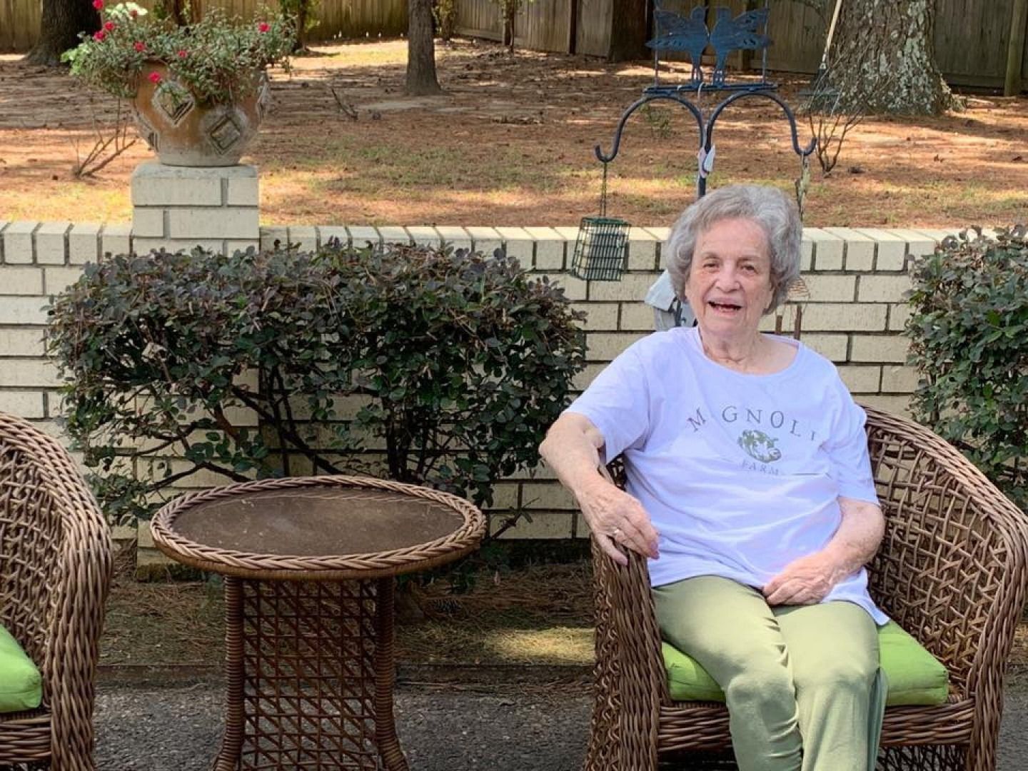 Elderly woman smiles in a wicker chair outdoors, near a small table and greenery.