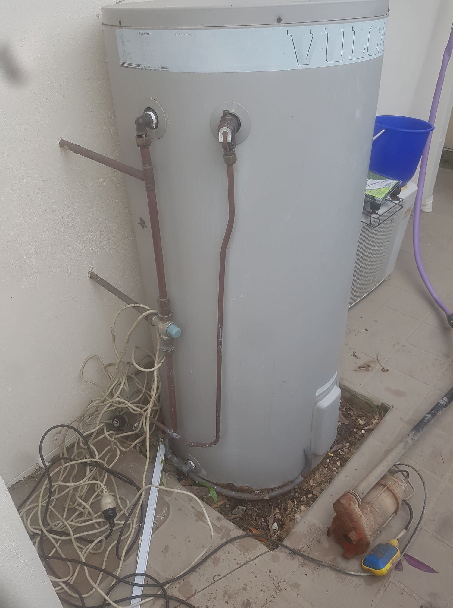 A water heater outdoors with pipes, wires, and a float switch against a wall. — 24/7 Hot Water in Valdora, QLD