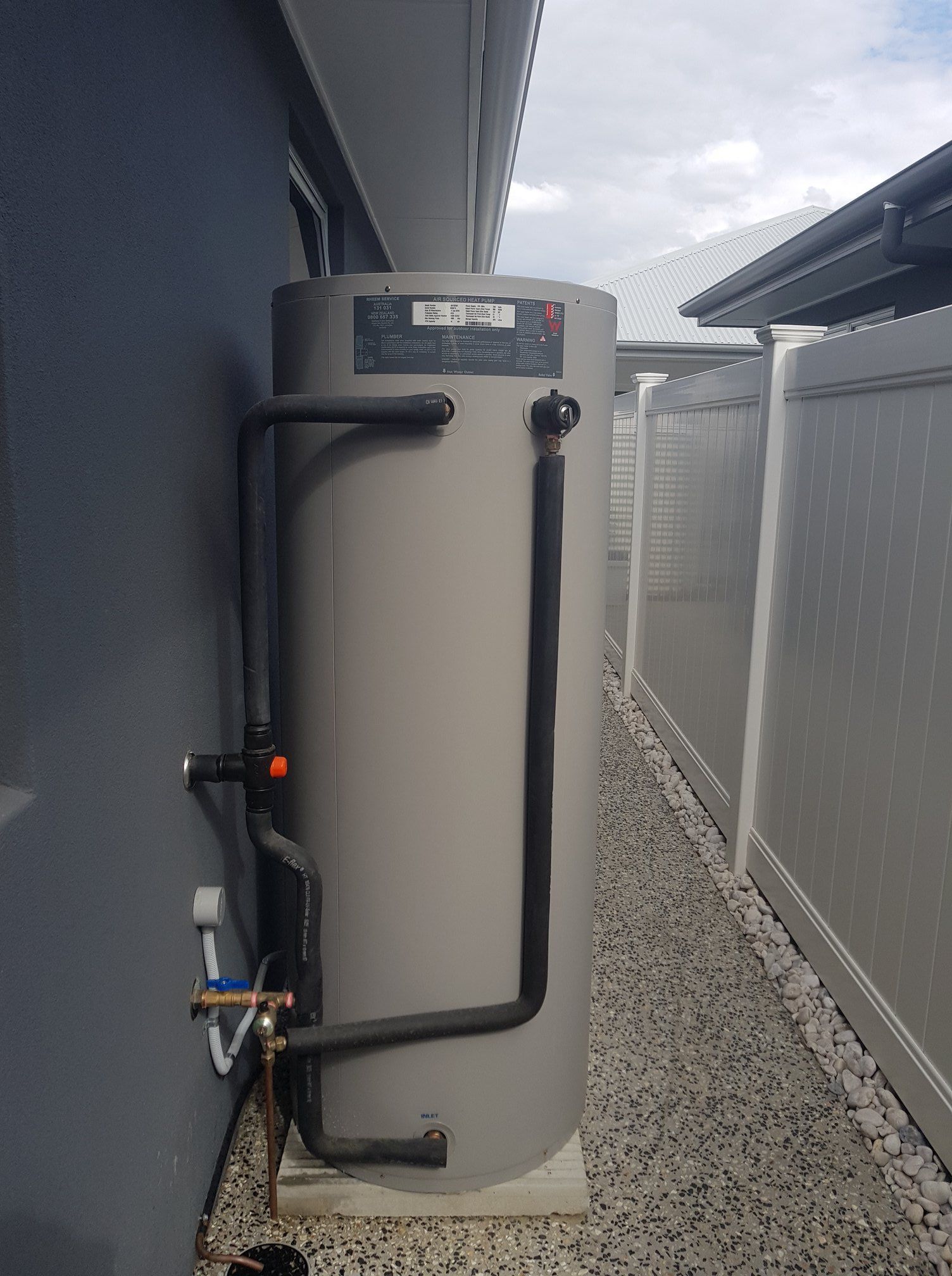 A Cylindrical Water Heater Next to a Gray Wall and Narrow Pathway, With Black Pipes — 24/7 Hot Water in Gympie, QLD
