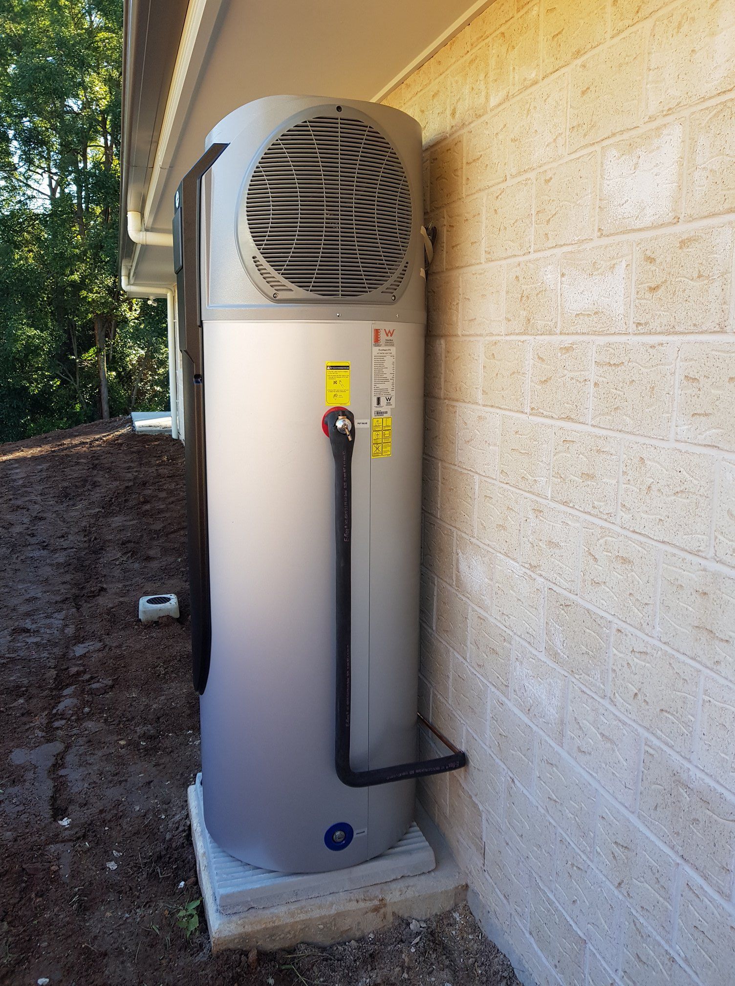 A Tall, Gray Heat Pump Water Heater Unit Installed on a Concrete Base Next to a Brick Wall — 24/7 Hot Water in Valdora, QLD