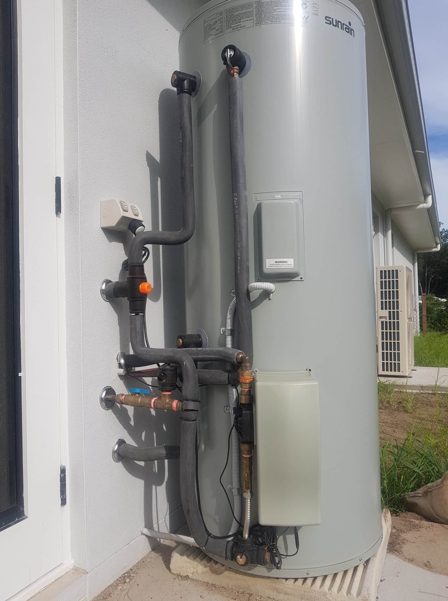 Large gray water heater mounted on a building's exterior wall with various pipes and fittings. — 24/7 Hot Water in Valdora, QLD