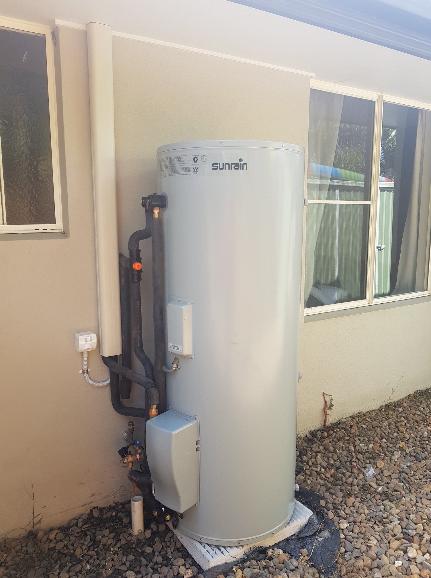 A Tall, Cylindrical Water Heater Against a Beige Building Wall — 24/7 Hot Water in Valdora, QLD