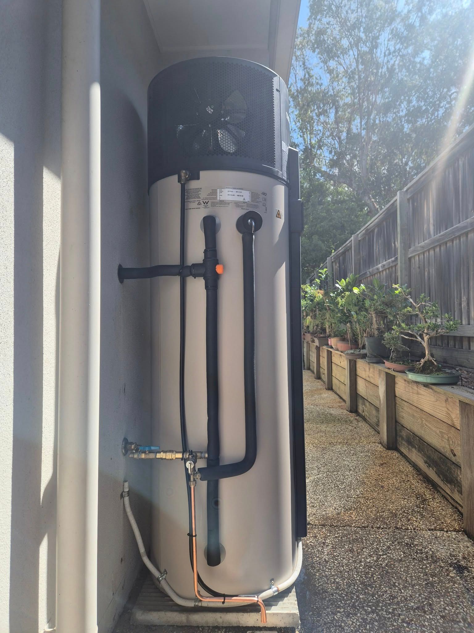 Tall Water Heater Against a Wall, Next to a Path With a Wooden Fence and Plants — 24/7 Hot Water in Valdora, QLD