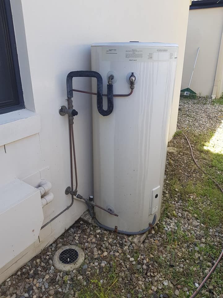 Exterior shot of a vertical water heater against a white building. Copper pipes and a black vent are visible. — 24/7 Hot Water in Valdora, QLD