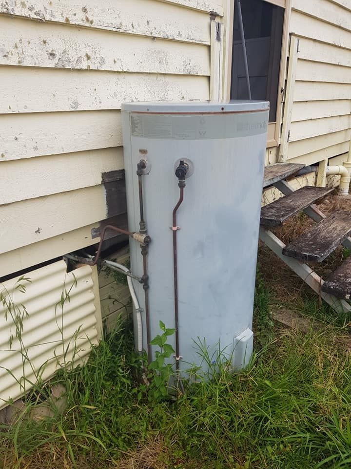 A tall, cylindrical hot water heater next to a weathered house exterior and wooden steps. — 24/7 Hot Water in Valdora, QLD