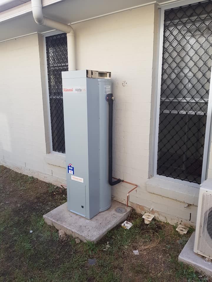 A Tall, Gray Water Heater Sits Outside Against a Beige Building Wall, Next to a Window — 24/7 Hot Water in Valdora, QLD