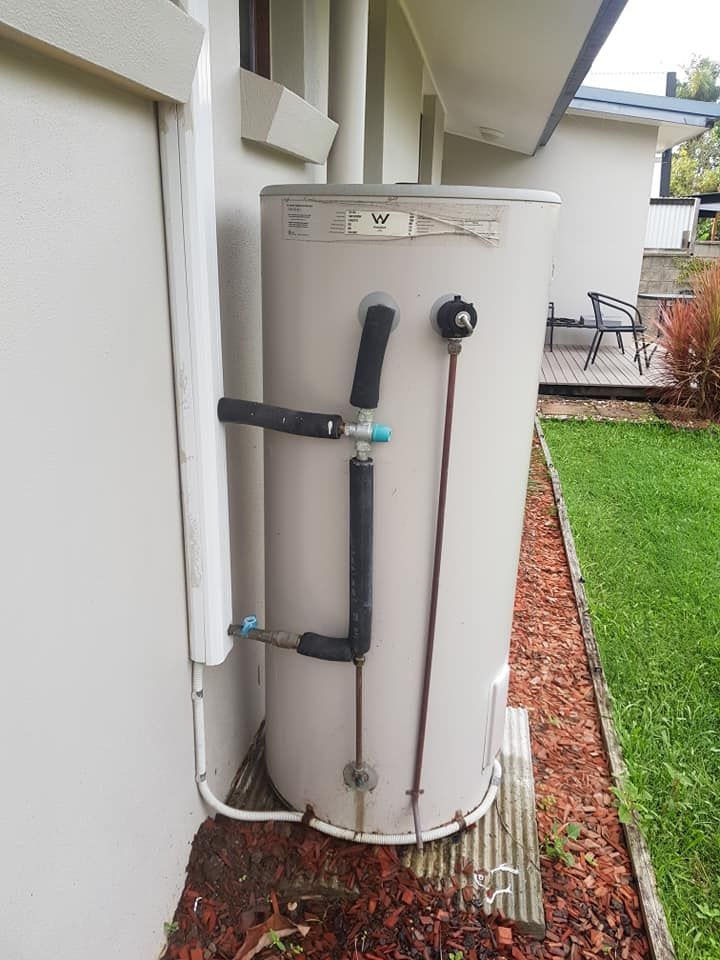 A tall, cylindrical water heater against a beige wall, with black pipes and copper tubing. — 24/7 Hot Water in Gympie, QLD