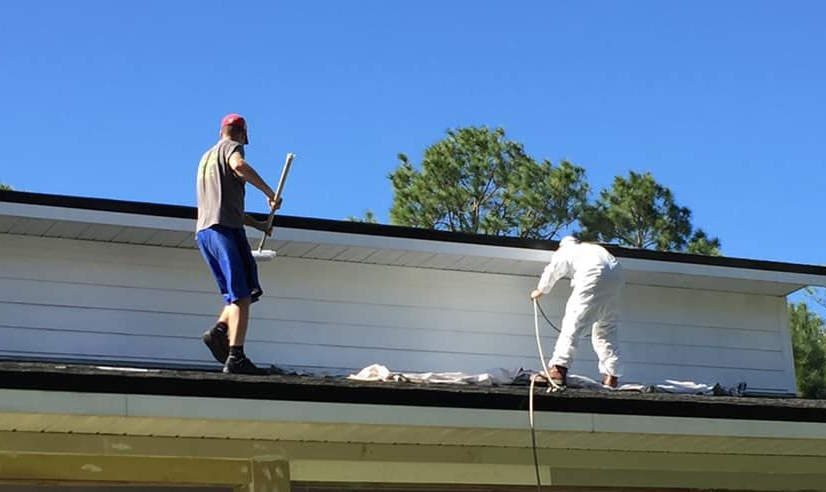 A hand uses a textured roller to apply tan paint to a rough, light-colored exterior wall.