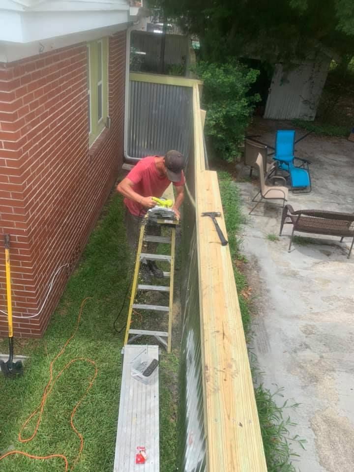 A person in a hat and grey shirt works on a wooden picket fence, using a level to ensure the top rail is straight.