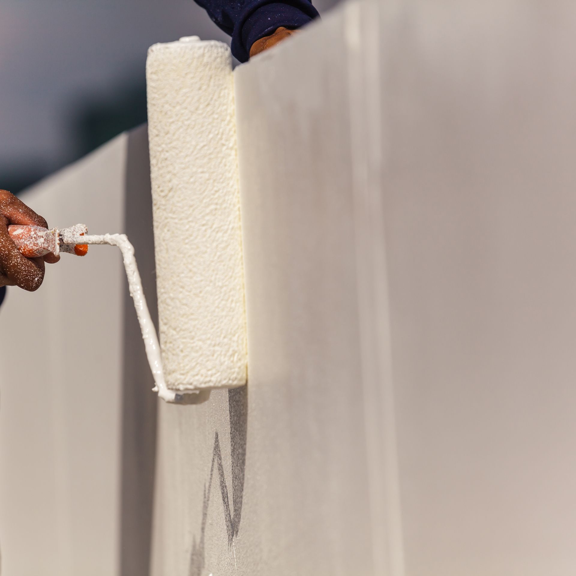 A person wearing orange rubber gloves uses a paint roller to apply white paint to a wall.