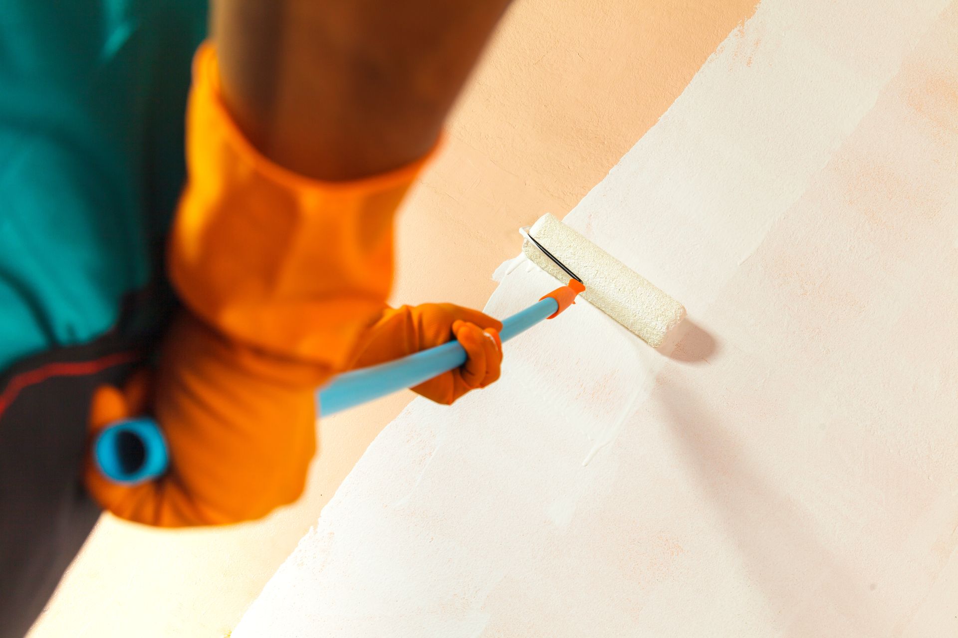 A worker in a yellow hard hat and blue uniform uses a paint roller to apply white paint to a grey wall.