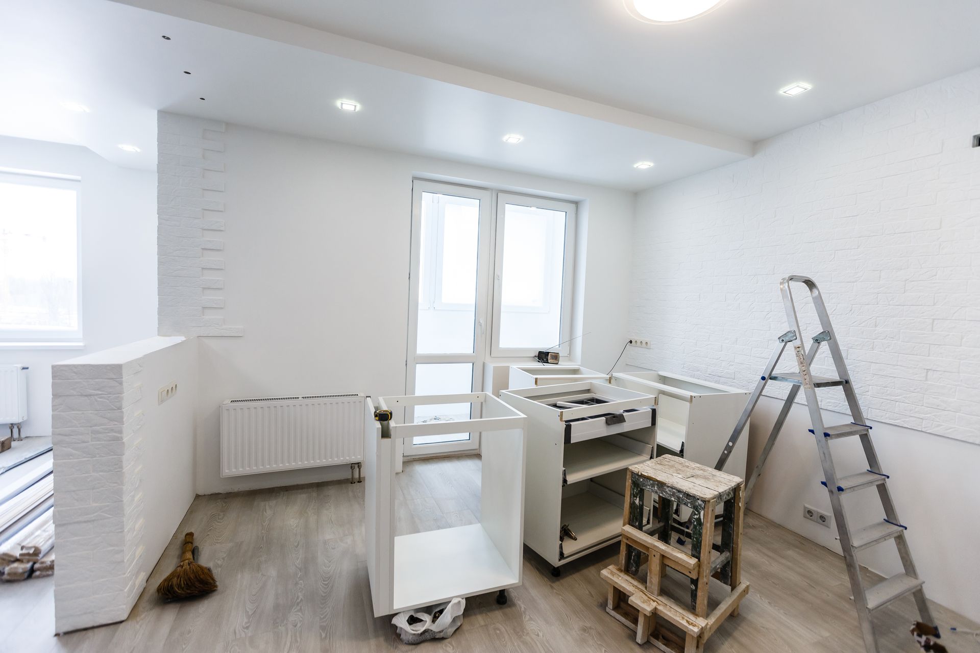 A brightly lit room under renovation with white walls, a metal ladder, and unassembled white kitchen cabinets on the floor.