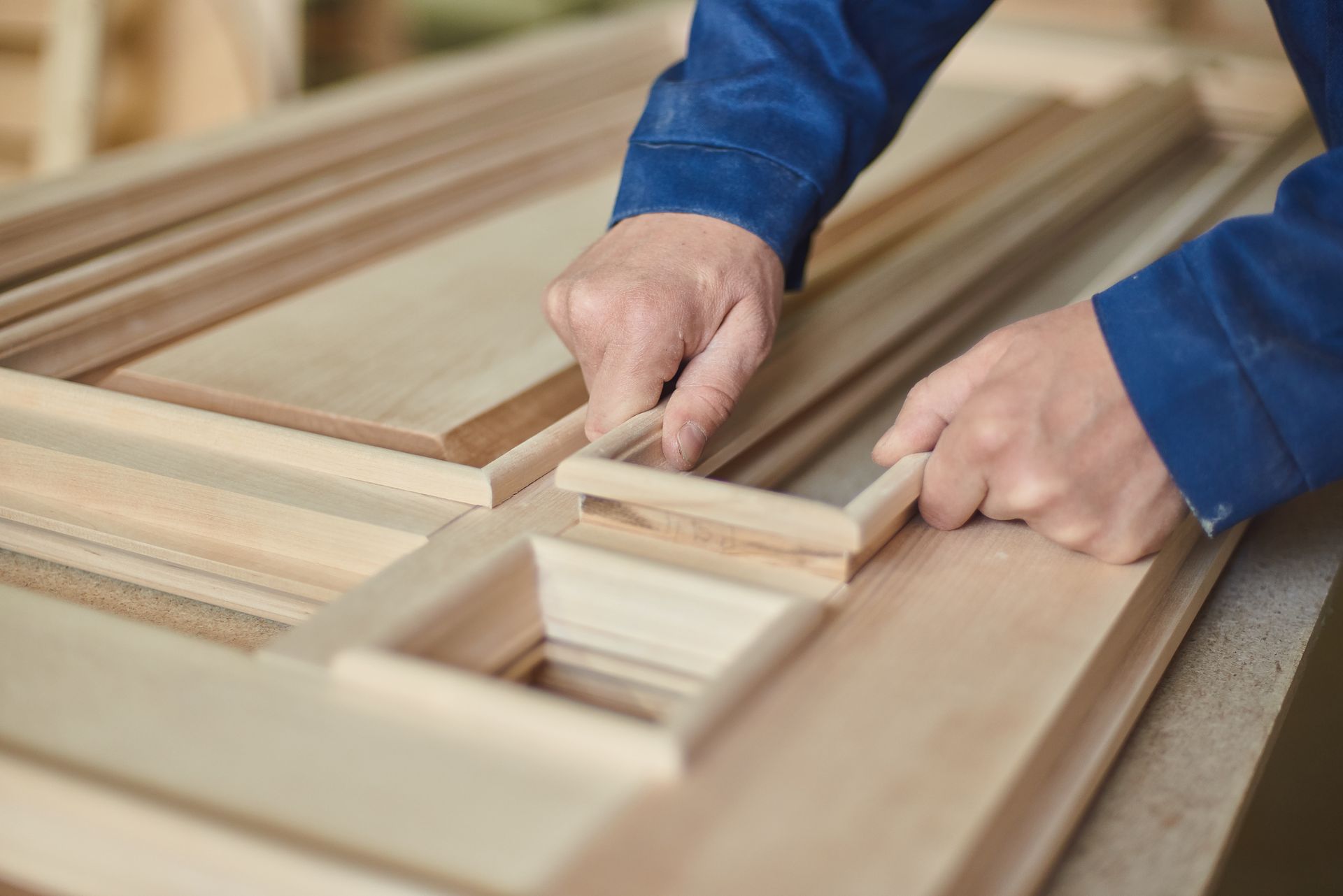 A person wearing a blue sleeve carefully attaches a thin wooden trim piece to a custom-made wooden door in a workshop.