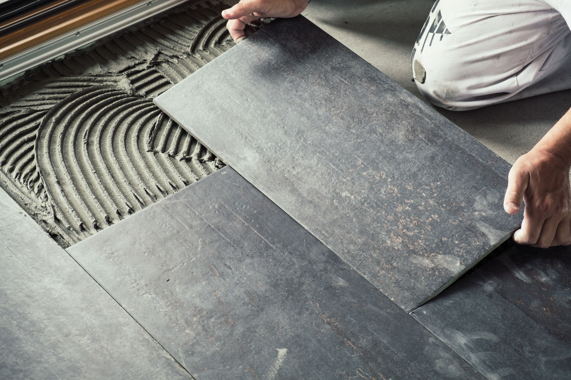A person placing a dark grey rectangular tile onto a floor with a layer of combed mortar adhesive.