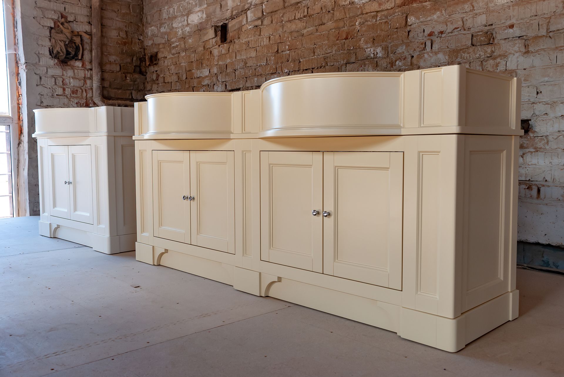 A pair of off-white double-basin bathroom vanity cabinets standing in a room with a rustic, unfinished brick wall.