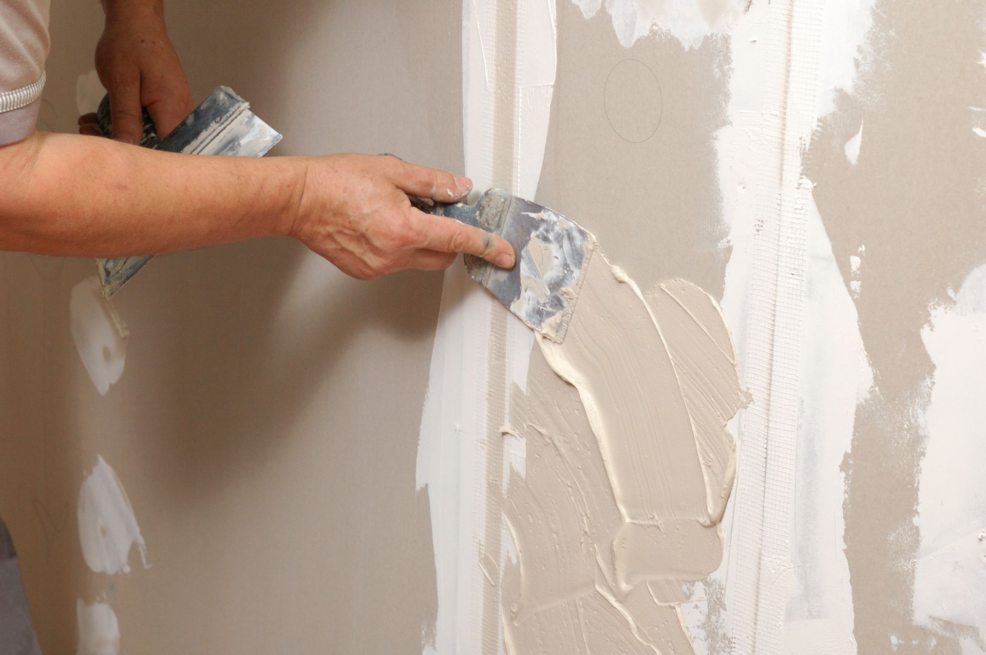 A close-up of a person using a metal putty knife to apply joint compound to a drywall seam on a wall.