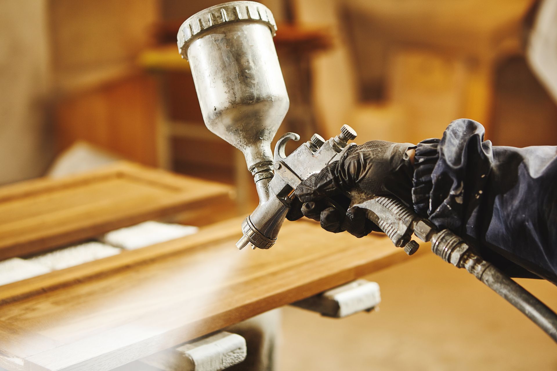 A close-up of a gloved hand using a pneumatic spray gun to apply a finish to a wooden board in a workshop.