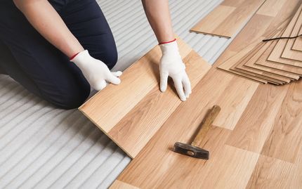 A person wearing white gloves installs light-colored wood laminate flooring over a gray underlayment with a hammer nearby. A person wearing white gloves installs light-colored wood laminate flooring over a gray underlayment with a hammer nearby.