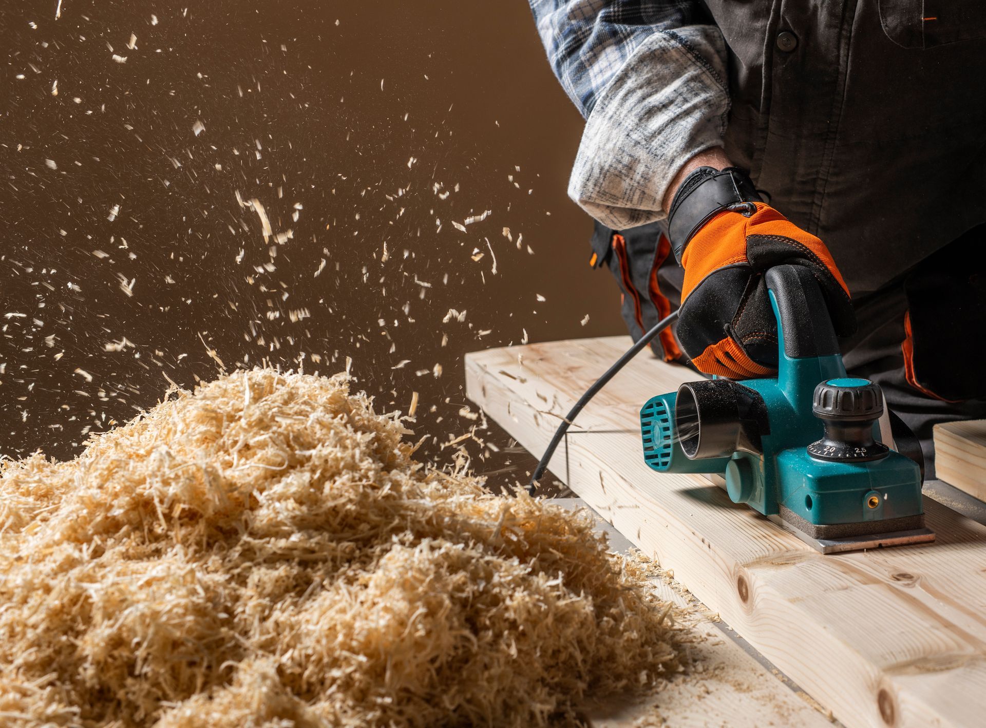 A woodworker uses an electric power planer on a wooden board, creating a large pile of sawdust shavings.