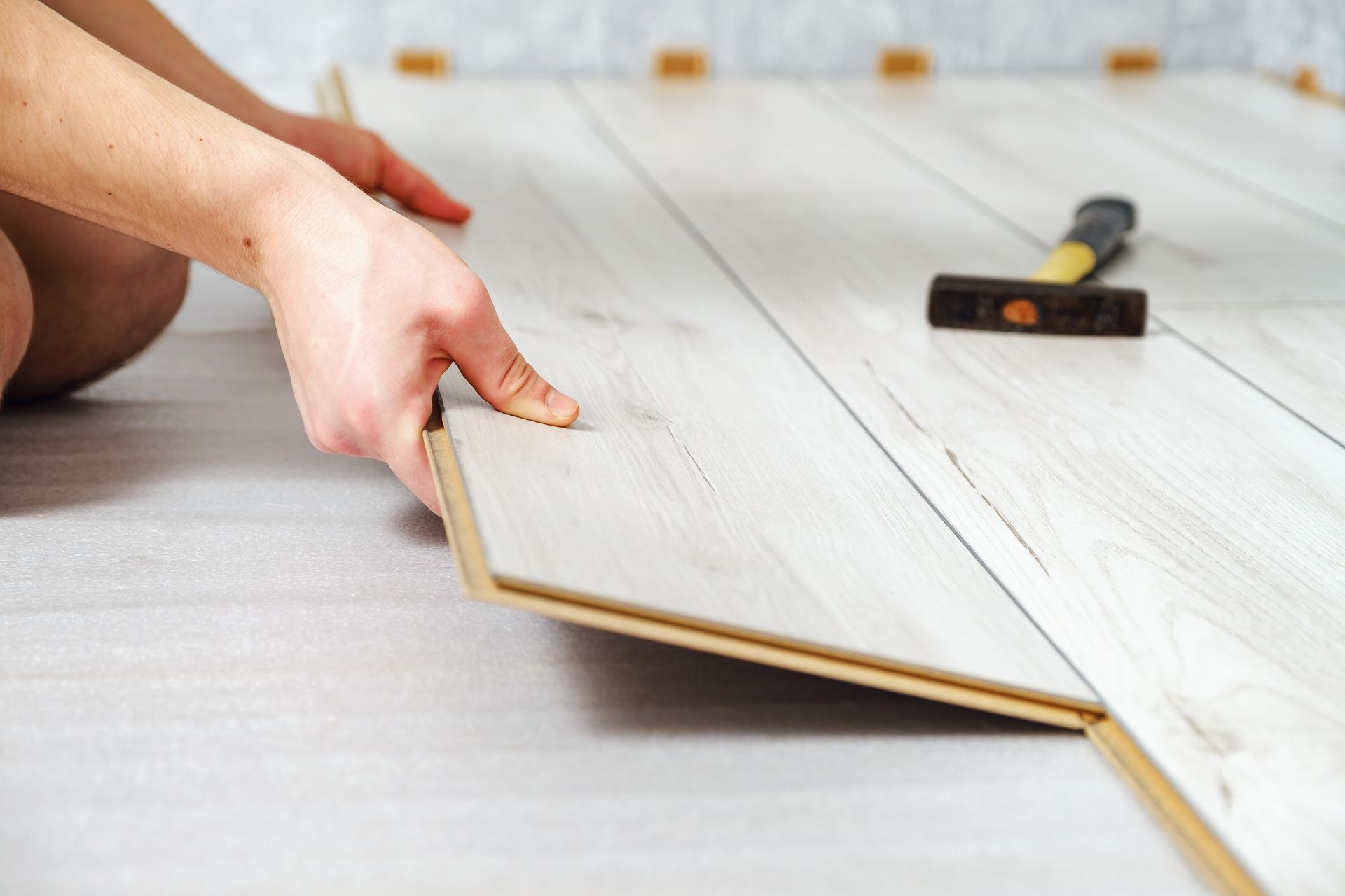 A pair of hands installs light-colored wood laminate flooring with a hammer resting on the planks nearby.