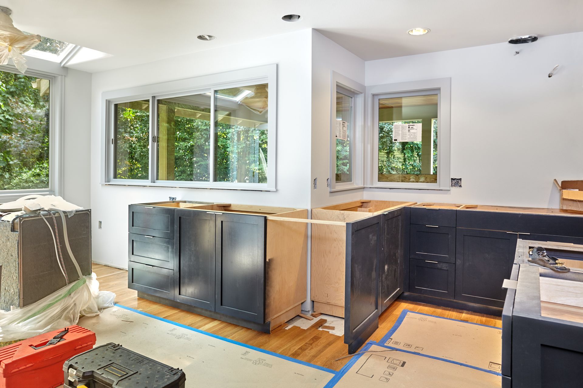 Kitchen under construction with dark blue cabinets installed beneath windows in a brightly lit, unfurnished room.