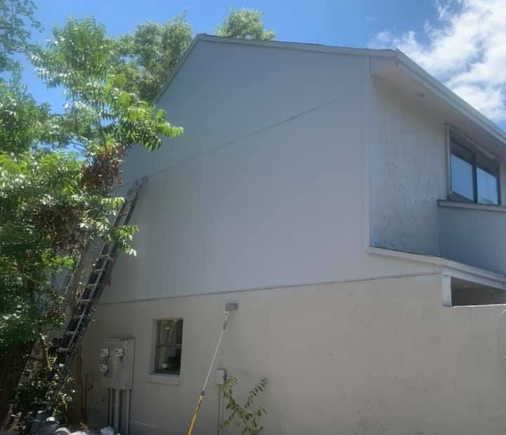 A worker uses a spray gun to paint a brick chimney and roof tiles blue against a bright, cloudy sky.