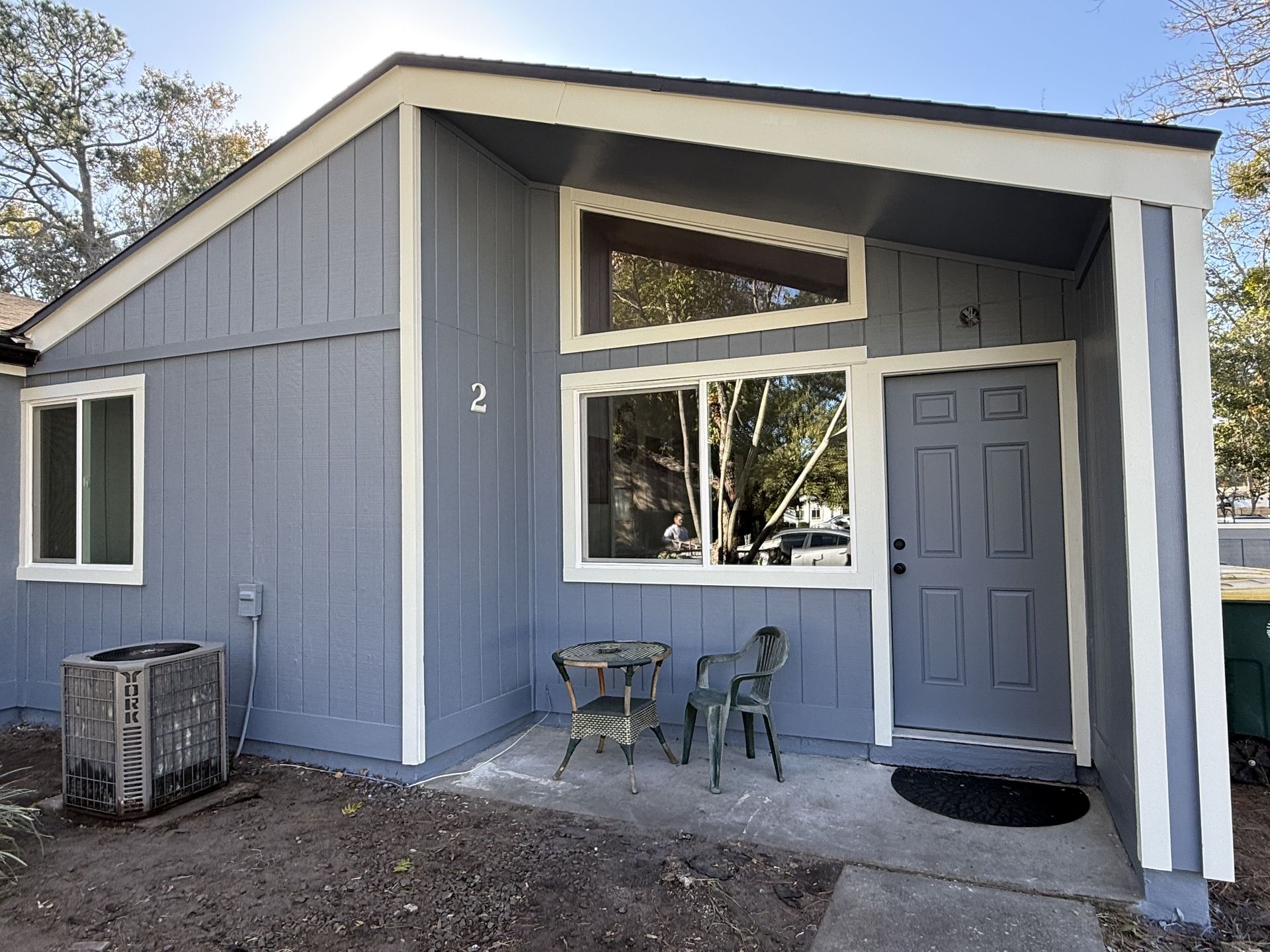 A low-angle view of a sage green, multi-story house with board-and-batten siding against a bright blue sky.